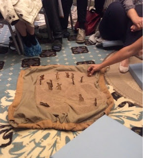There is a cloth on the flloor covered in sand. Little wooden figures representing people have been placed in the sand, and a hand is reaching out to place yet another.