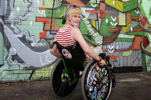 Young woman with blonde, short hair, wearing sunglasses up on her head, in a wheelchair decorated with myriad stickers, in front of a graffiti-covered outdoor wall.