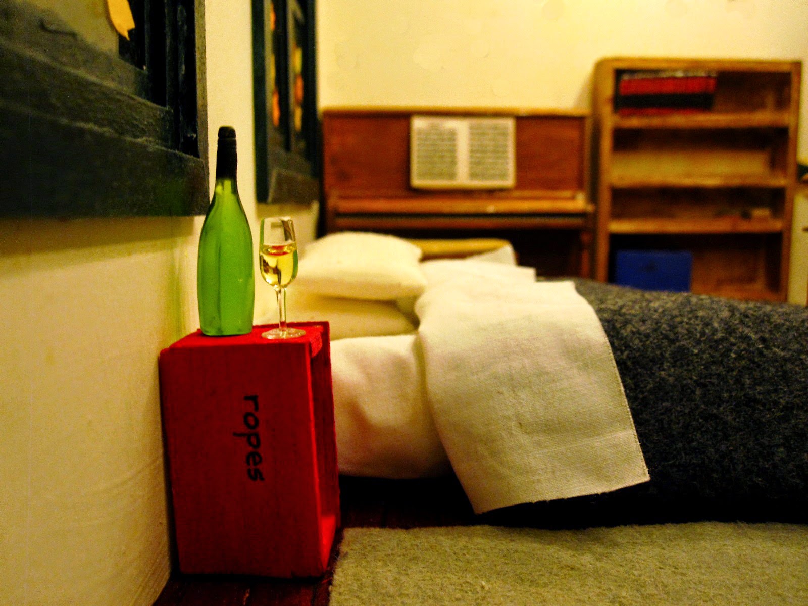 Miniature scene with a mattress and bedding on the floor, a red crate marked 'ropes' next to it with a glass and bottle of wine on it, and a piano in the background.
