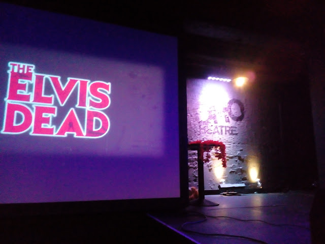 The stage before The Elvis Dead show opened: the logo projected on a screen next the stage, which is empty except for a small table with a few props.