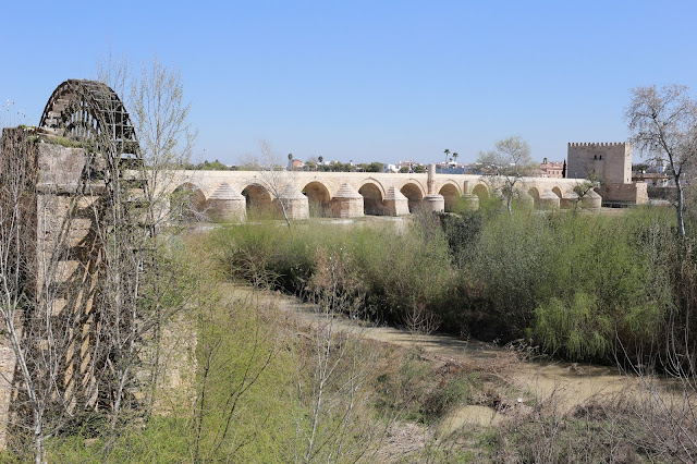 Antigua rueda de madera hidráulica en un cauce seco y el puente romano de Córdoba y el rio Guadalquivir al fondo.