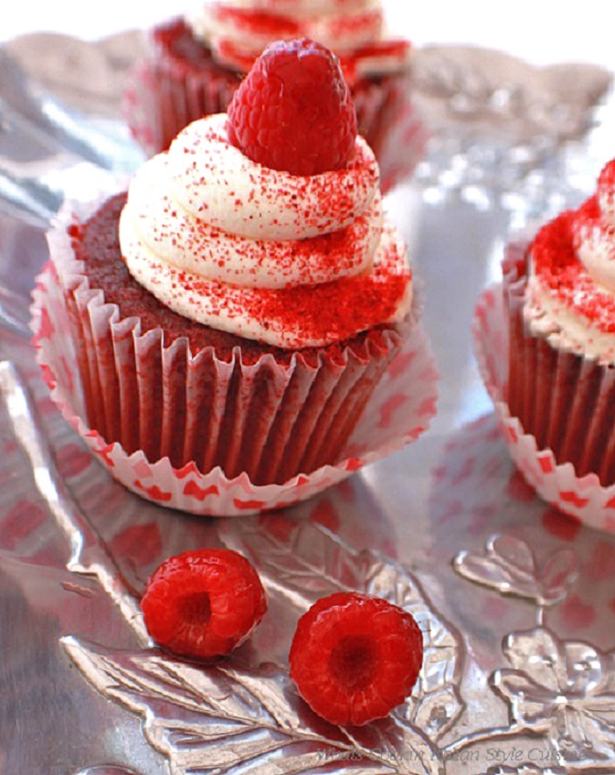 this photo is of two cupcakes that are raspberry and red velvet. They have whole raspberries on top and cream cheese frosting in heart paper liners