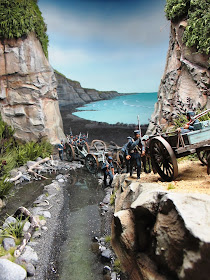 Diorama of 19th-century soldiers crossing a creek and mounting a track by the sea.
