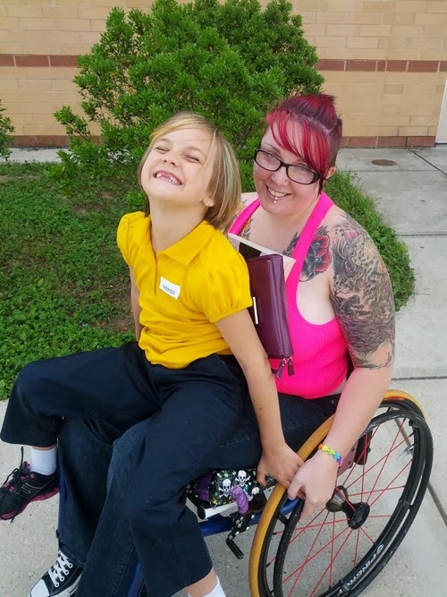 Woman with pink top and glasses in wheechair, with young girl sitting in her lap, both smiling