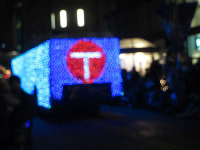 Blurry photo of a bus wrapped in blue Christmas lights with a red circle on the back with a white T in it