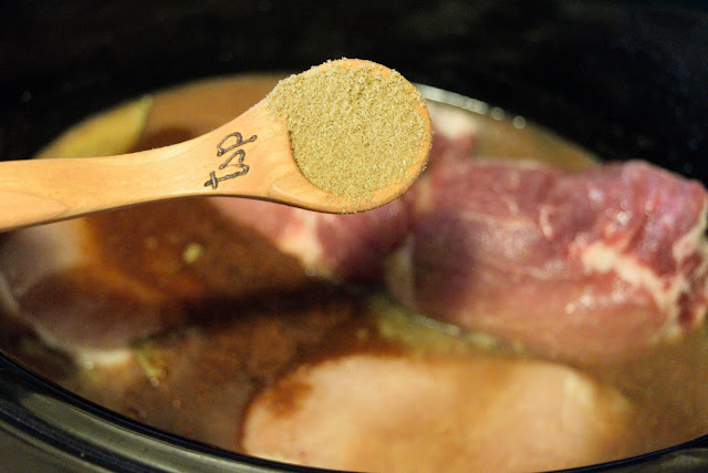 A measuring spoon with cumin in it, over the slow cooker with the pork in it. 