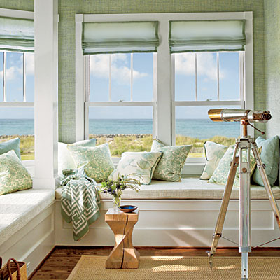 Window seat vignette with an ocean view in Coastal Living Magazine showhouse with wood floor, a seagrass area rug, a telescop, mint sea grass wallpaper, Mint roman curtains on casement windows, and a COCOCOZY Logo Throw