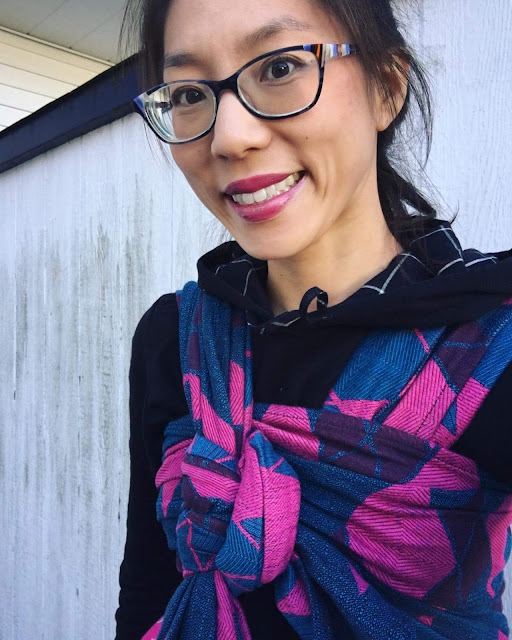 [Image of a smiling tan skin bespectacled Asian woman wearing a not-visible toddler on her back in a pink and blue large argyle patterned woven wrap carrier over a black sweatshirt with a plaid-lined hood. They’re outside in front of a worn white shed.]