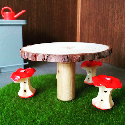 Three 1/12 scale modern miniature stools in the shape of apple cores around a table made with a slice of tree with bark still on it, on a round mat of fake grass. In the background is a bar with a red watering can on it.