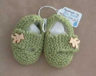 An above view of a pair of knitted green baby shoes made by Kelly Hickman. The label is still attached. Each shoe has a wooden button in the shape of a teddy bear. The shoes are stuffed with white cotton wool to keep their shape.