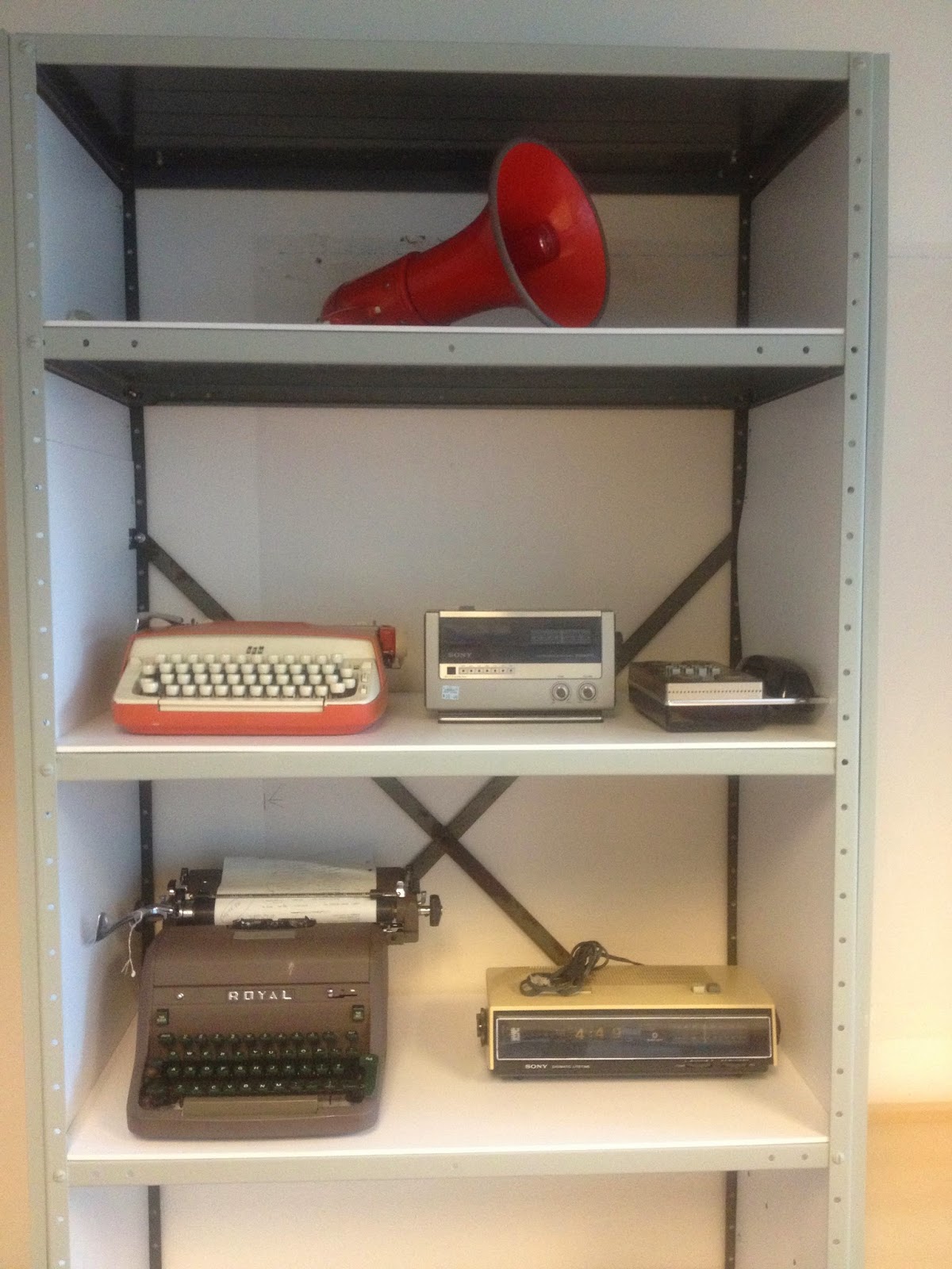 Collected artifacts on shelves from the late seventies. A red bullhorn, two typewriters, a radio, clock, and dial pad phone sit on the shelves.