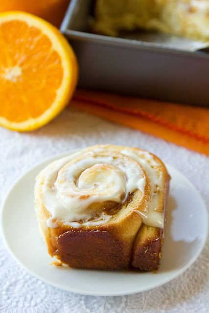 An orange marmalade cinnamon roll sitting in front of a tin of rolls and an orange