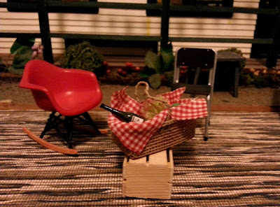 Two modern dolls' house miniature chairs and a picnic basket full of food, on a dining table in front of a dolls' house miniature school.