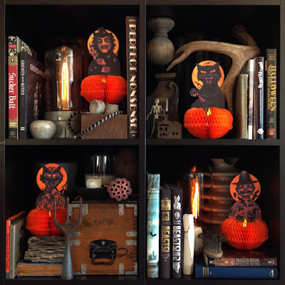 Honeycomb decorations (witch, scarecrow, black cat, and devil) on a shelf with various creepy and spooky books. 