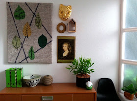 One-twelfth scale modern miniature office scene containing a credenza with a black Eames chair next to it and an artwork with leaves above it. On the credenza are three green lever-arch folders, a bowl, a vase, a potted plant and a ceramic ornament.