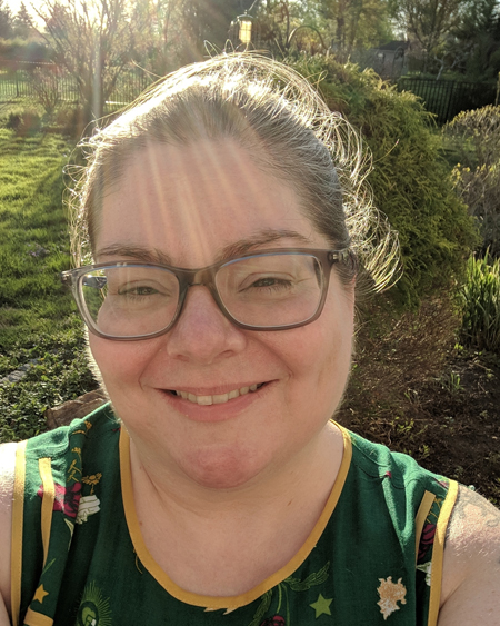 image of me from the shoulders up, wearing a green tank-top and grey-framed glasses, with my hair pulled up, standing in my garden with sun rays from the setting sun behind me