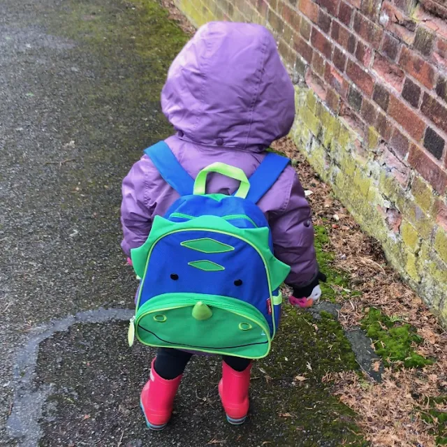 Back view of a toddler wearing the Skip Hop Zoo Dinosaur backpack, a purple coat and pink wellies