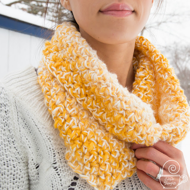Image of a tan skin Asian woman from smile through top torso featuring a yellow and cream crocheted cowl worn with a white sweater. In the background is a worn white shed adn winter wonderland.