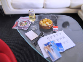 Modern dolls' house miniature Noguchi coffee table holding a glass of champagne, a bowl of chips, a Virgin Australia magazine, a selection of postcards, a pen and a sheet of address labels.