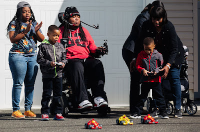 a family of five playing with r/c cars. Two of the cars are being controlled via remote control by two able bodied boys. The third is being controlled by a man in a wheelchair with a headset that has a pipe. Sipping and puffing into the pipe controls the r/c car.
