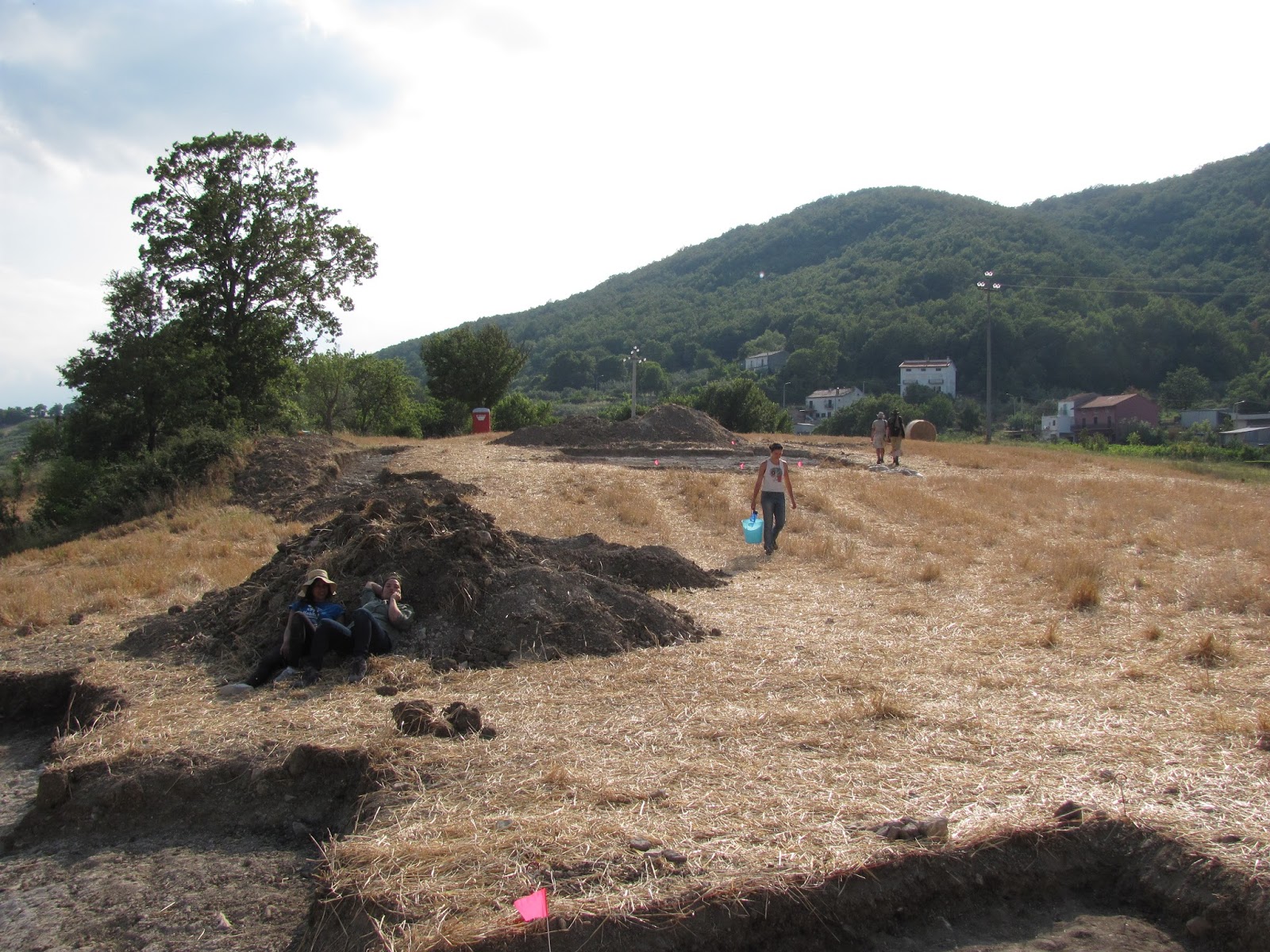 On a hillside, two people rest against a pile of dirt while another carries a bucket. A wide, shallow area is dug out.