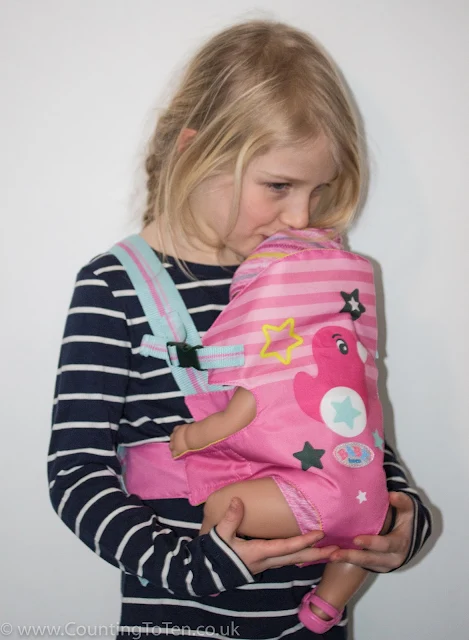 A girl kissing her BABY born dolls head while wearing her in a pin and blue BABY born carrier seat