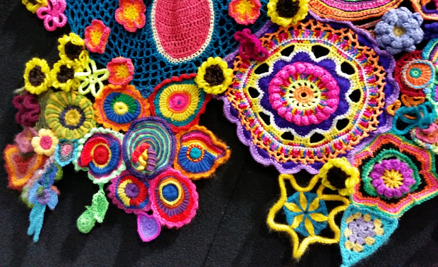 The lower left hand section of the second panel adjacent to a giant pink flower. The large, white-bordered, pink petal of that flower is along the top edge and surrounded by blue crochet mesh.