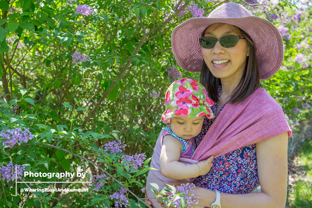 [Image of me, a smiling light tan skin Asian woman with dark brown hair, sunglasses, and a floppy purple sun hat in front of a lilac bush in bloom. I am wearing a toddler on my hip. She has on a floral sun hat and is contemplating picking the purple lilacs off the bush. The sweet fragrance wafts into the spring air with every warm breeze. We are using a pinkish reddish linen woven wrap, in a carry called poppins hip carry with the shoulder spread out. The darker side and lighter side of the woven wrap are both visible.]