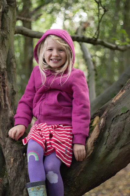 A girl in winter clothes: thick cardigan, tights and wellies sitting in a tree