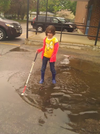Girl in puddle, wearing rainboots with wet hair and white cane.