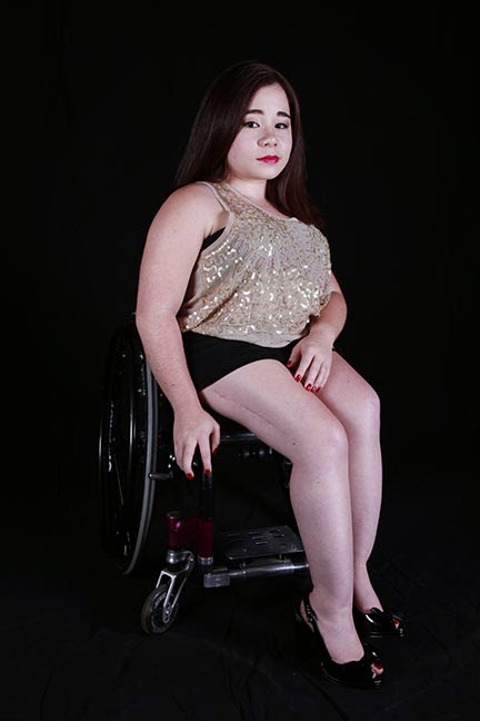 A picture of a young female with a gazing expression, wearing a sparkle gold tank top and a black skirt, sitting in a purple wheelchair, with a black background. Her legs are angled outside of the chair and a scar appears on the right side of her thigh.