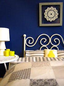 One-twelfth scale modern miniature bedroom with deep blue wall, white wire bed and side table and grey and white bedding.