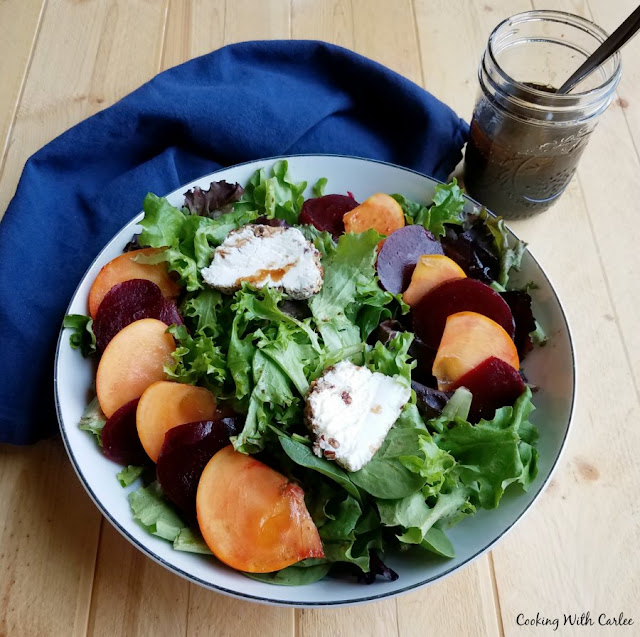 A fun sweet and savory salad with beets, persimmons, goat cheese and pecans lightly dressed in a homemade salad dressing. It is a perfect starter or a great lunch. greens topped with sliced beets, persimmons and goat cheese with jar of dressing