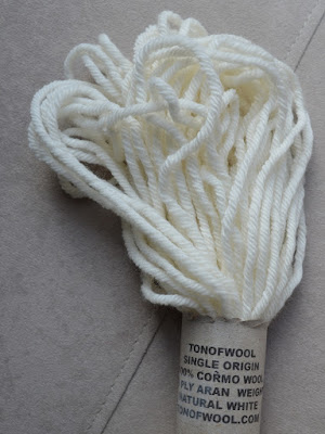 A close-up photo of one half of the skein with the paper label which says: TONOFWOOL SINGLE ORIGIN 100% CORMO WOOL 10 PLY ARAN WEIGHT NATURAL WHITE TONOFWOOL.COM