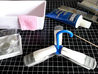 One-twelfth scale modern miniature bath with pink towel over the side, with a floor-standing 'tap' on top of a small round silver tin, with clamps holding it in place. In the background is a tube of glue.