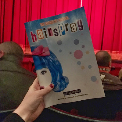 a hand holding up a blue Hairspray programme in front of red theatre curtains