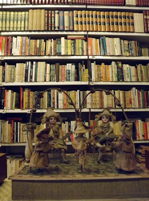 Uma prateleira de livros ao fundo e uns bonecos bailarinos de cerâmica