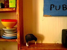 Dolls house miniature wall shelving with a kiwi ornament placed on it, next to a pile of plates and dishes  and a shelf of food items.