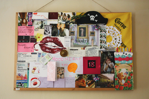 a noticeboard full of photographs, handwritten notes, birthday cards, travel parephenalia, post it notes, paintings, and various trinkets and gifts
