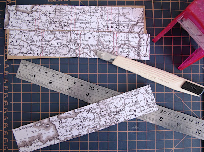 Pieces of scrapbooking paper showing a vintage map of France laid out on a cutting matt with a metal ruler, a knife and a sticker machine.