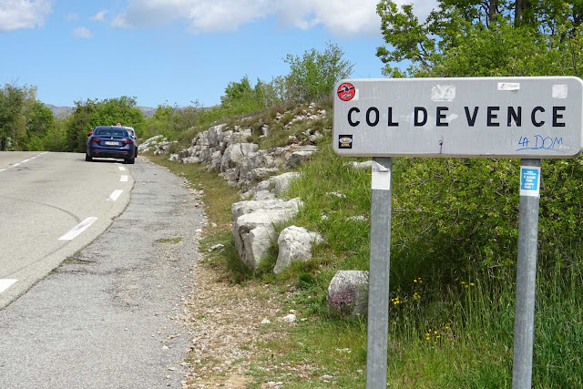 Schild mit Col de Vence, blaues Auto, Gräser und Steine
