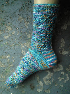 Someone wearing a knit sock.  The sock is blue with flecks of other colours, and knit in a stretchy ribbed lace.