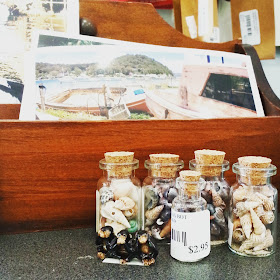 Selection of bottles containing miniature shells in front of a box displaying postcards.