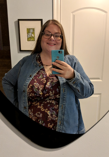 image of me standing in a hallway mirror, pictured from the waist up, with my hair down, smiling, wearing grey-framed glasses, a dark pearl necklace, a burgundy blouse with a floral pattern, and a medium wash collarless denim jacket