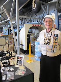 A woman dressed in a 1940s outfit, standing in front of her stall at a miniatures convention.