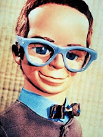 TV puppet character, wearing blue, glasses, blue shirt and black and polka dot blue bow tie,