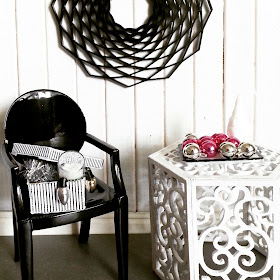 Modern miniature scene of a black ghost chair next to a white scrollwork side table. On the chair is a shoebox filled with Christmas decorations, and on the table, a tray displaying Christmas baubles in  silver and hot pink.