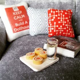 Close-up of a one-twelfth scale modern miniature sectional sofa in grey velvet. On the sofa are three cushions in red and pale blue, a plate of cakes with a glass of water on it, and a book.