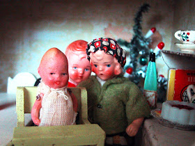 Three vintage dolls-house dolls pose for a picture in front of a Christmas tree and a sideboard set up for Christmas