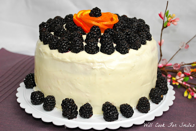 whole cake on a cake platter with white frosting and blackberries all around the bottom and blackberries all over the top with a orange peel made to look like a flower with a blackberry in the middle 